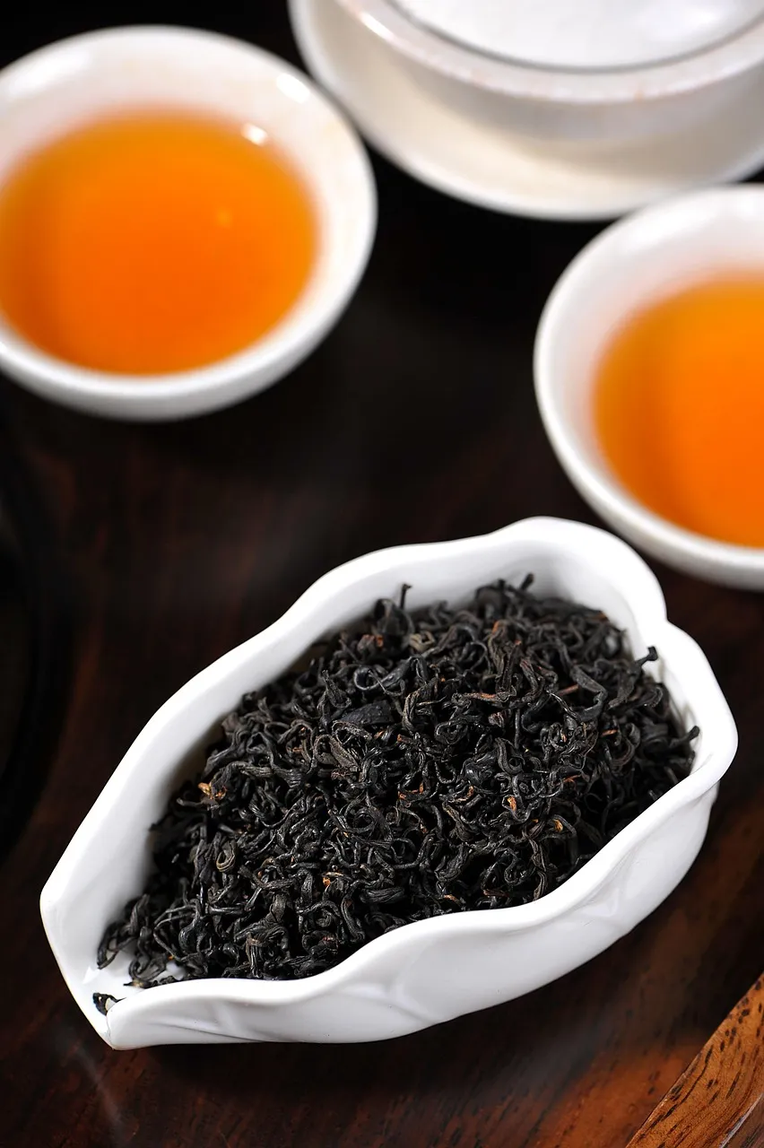 Des feuilles de thé noir torsadées dans un bol à côté de tasses de thé rouge ambré