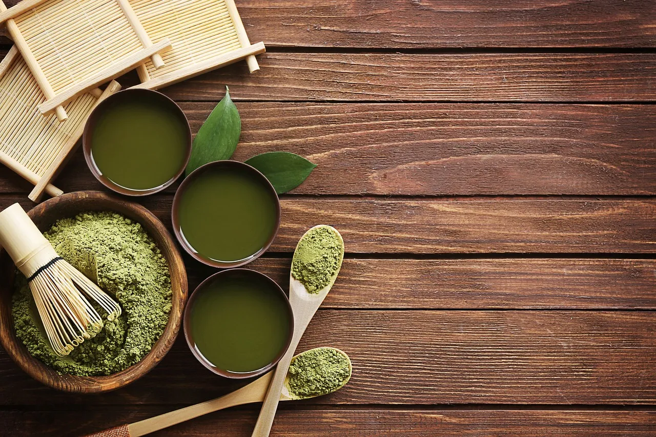 De la poudre de matcha, un fouet en bambou et des tasses de matcha battu sur une table en bois